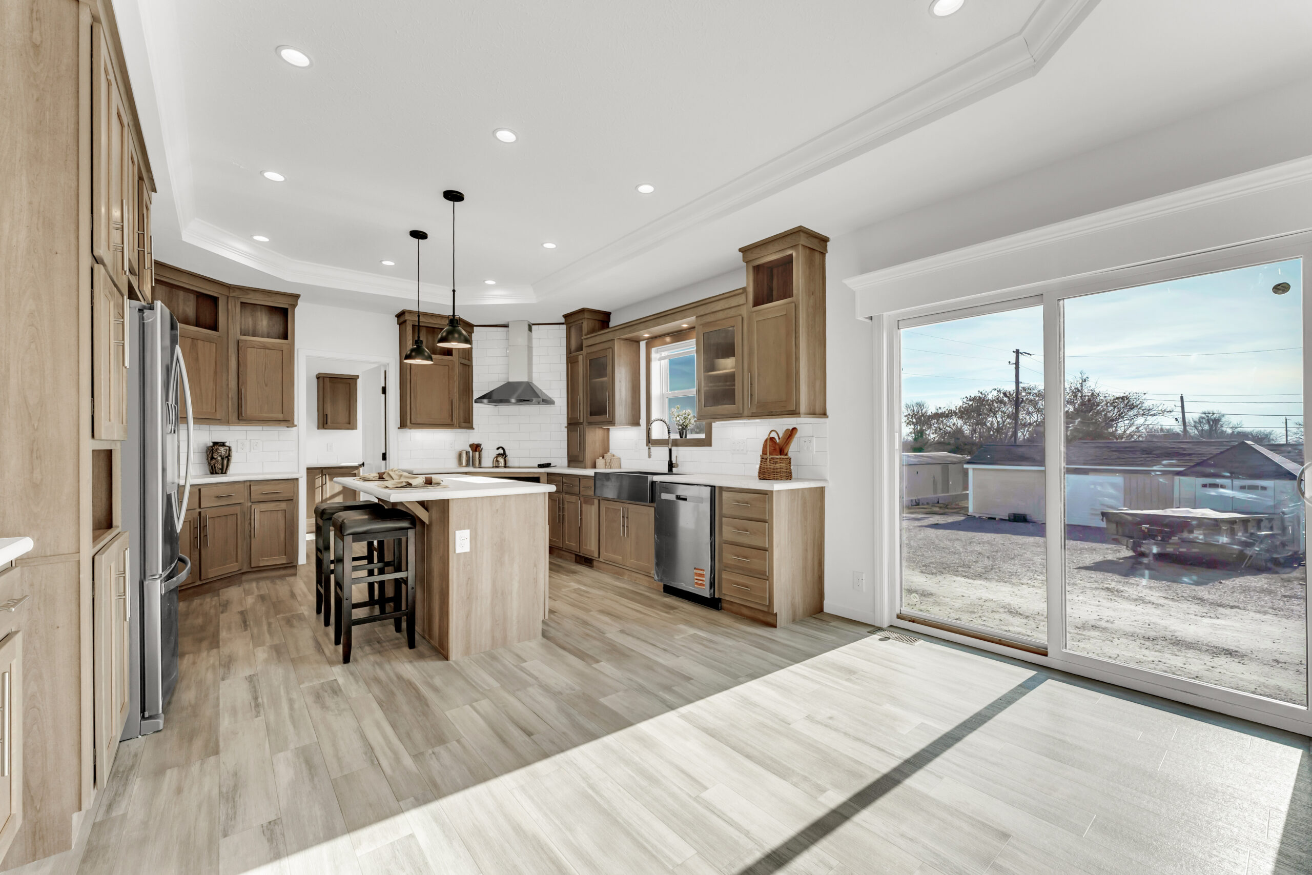 Spacious kitchen with natural wood cabinets and a large island. Pendant lights hang above, and a sliding glass door offers views of a sunny backyard.