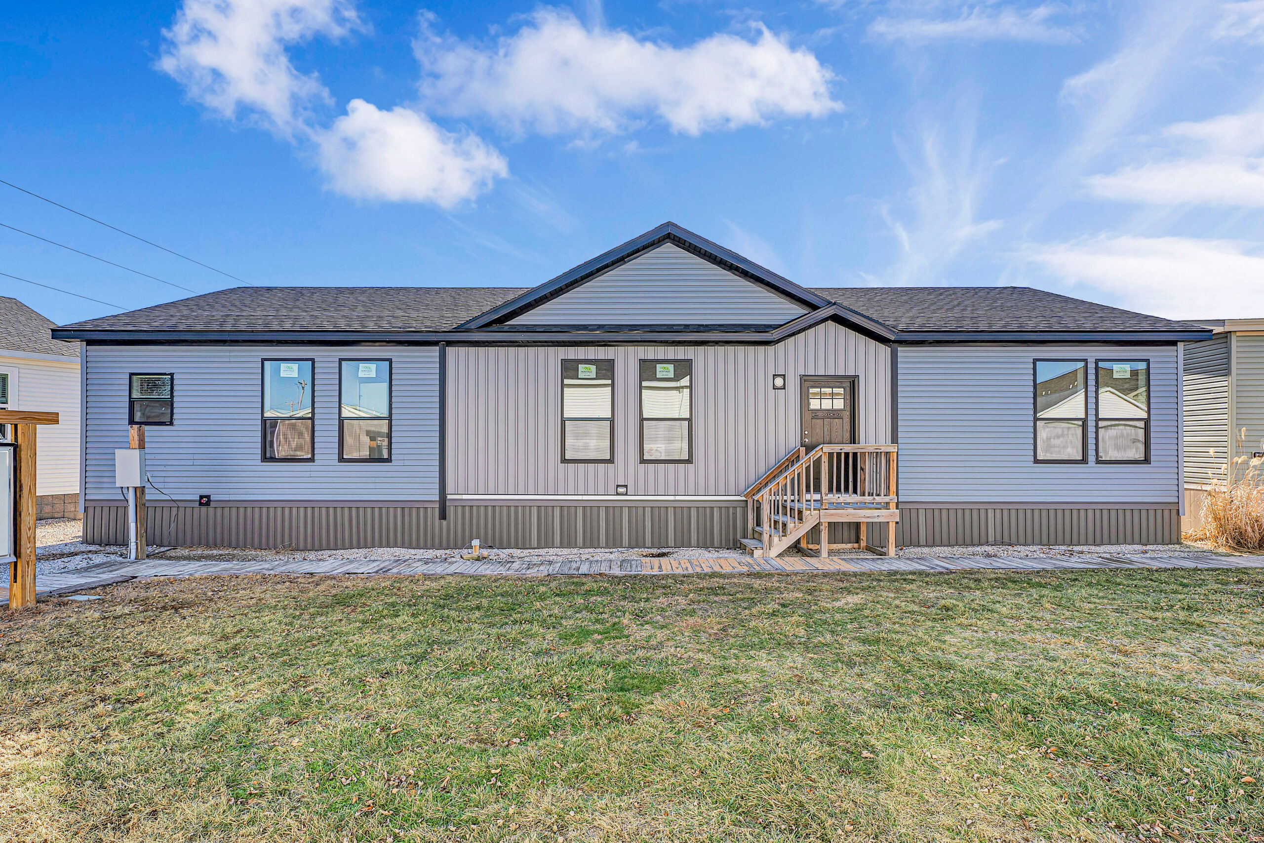 A modern, single-story home with gray siding, black-trimmed windows, and a small wooden porch. The house sits on a grassy yard under a blue sky.