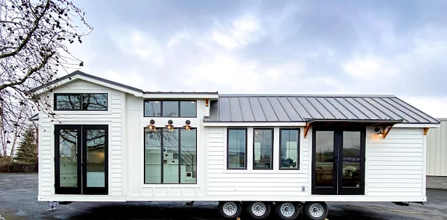 Modern tiny house on wheels with white siding, large windows, metal roof, and black-framed glass doors. Set against a cloudy sky, it conveys a sense of mobility and minimalist living.