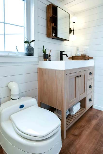 Compact bathroom with a composting toilet, wooden vanity with a white sink, black faucet, round mirror, and small plants. Bright, clean, and minimalistic.