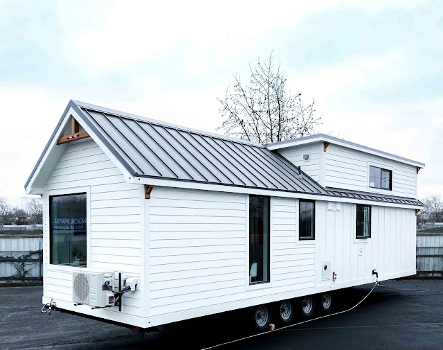 A modern, white tiny house on wheels with a sleek metal roof is parked on asphalt. It features large windows and a compact, minimalist design.