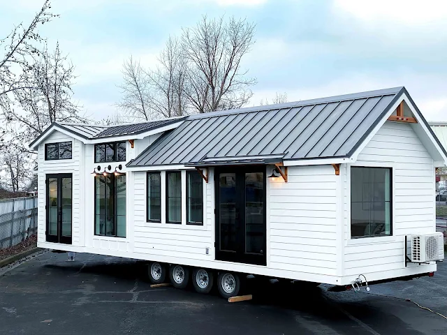 A modern white tiny house on a trailer, featuring large glass doors and windows, a metal roof, and a serene outdoor setting with bare trees.