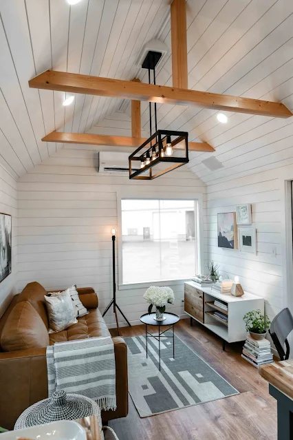Cozy living room with a vaulted ceiling features exposed wooden beams and modern pendant lighting. A brown leather sofa faces a small table with flowers, atop a geometric rug. Wall art and a cabinet add decor, creating a warm, inviting atmosphere.