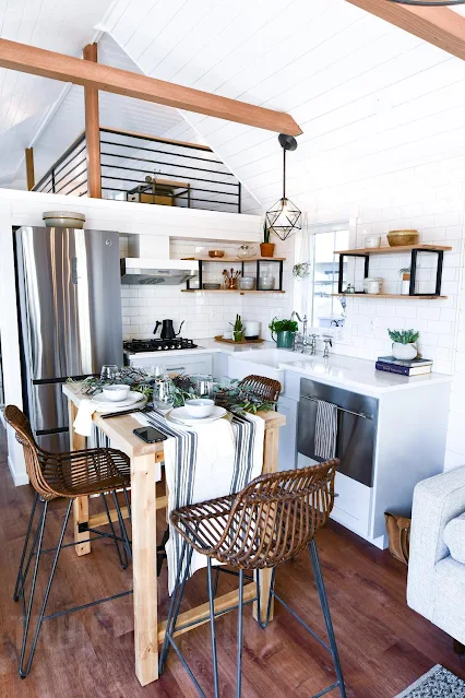 Bright, modern tiny kitchen with white walls, wooden floors, and a high ceiling. Features a dining table with wicker chairs, stainless steel appliances, and open shelves.