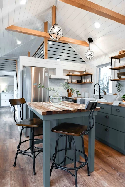 Airy kitchen with rustic charm, featuring a wooden island with metal stools, pendant lighting, green cabinets, and a stainless steel fridge. Cozy and inviting.
