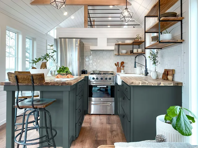 Modern kitchen with dark green island and light wood countertops, open shelving, stainless steel appliances, and geometric pendant lights.