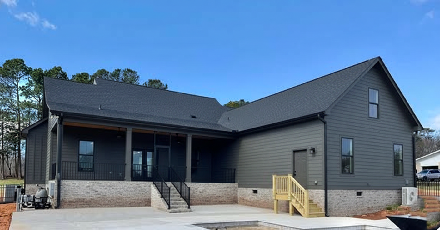 Modern gray house with a steep roof, large porch, and brick accents. A small wooden staircase leads to a patio. Bright blue sky and trees in the background.