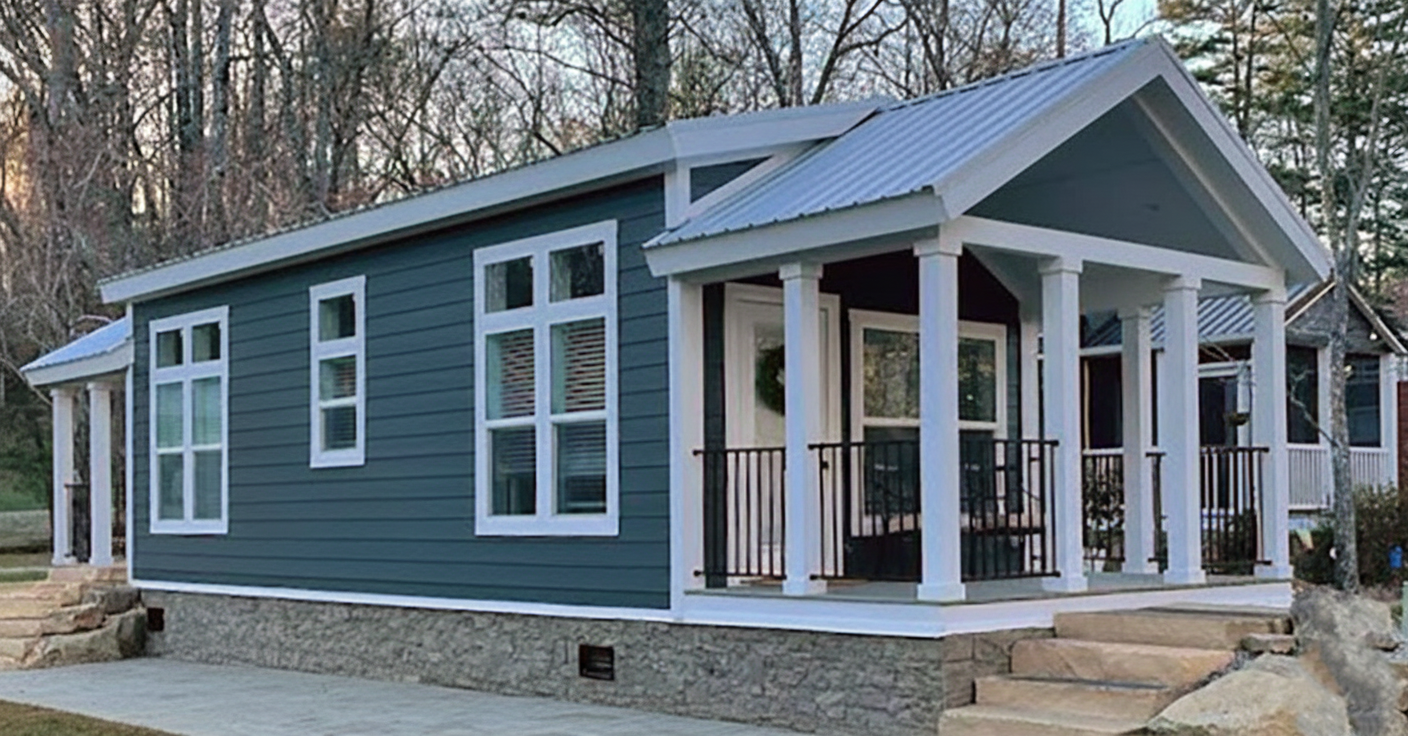 A modern tiny house with blue siding and a stone foundation, featuring a white-trimmed porch and large windows, set in a wooded area, conveying a cozy, peaceful vibe.