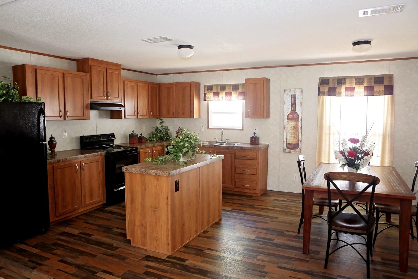 A warm kitchen with wooden cabinets and flooring. A dining table with flowers is by the window. The space feels cozy and inviting, accented by natural light.
