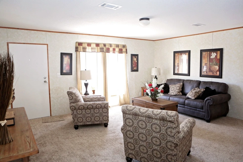 Cozy living room with a dark sofa, patterned armchairs, and a wooden coffee table adorned with flowers. Soft lighting, art pieces, and a large window create a warm atmosphere.