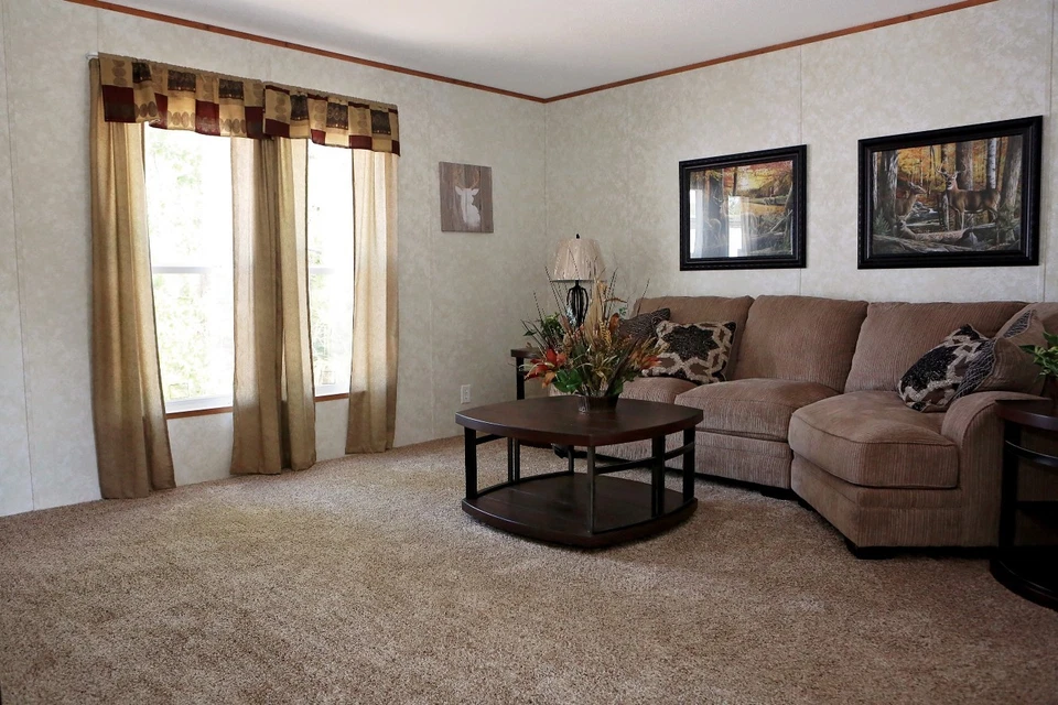 Cozy living room with a brown sectional sofa, decorative pillows, and a wooden coffee table with flowers. Cream curtains filter soft light, creating a warm atmosphere.