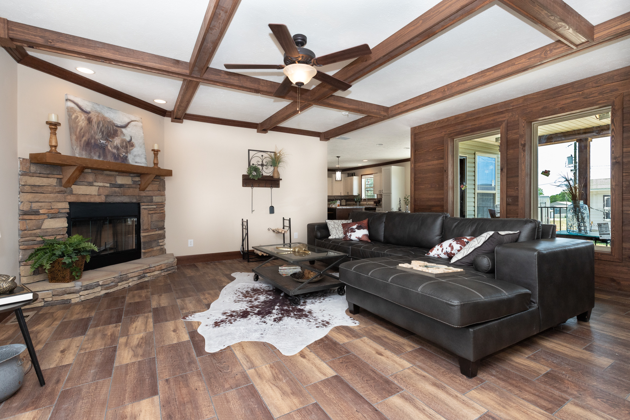 Spacious living room with cozy fireplace, dark leather sectional, and a wooden coffee table on a cowhide rug. Ceiling fan above and large windows.