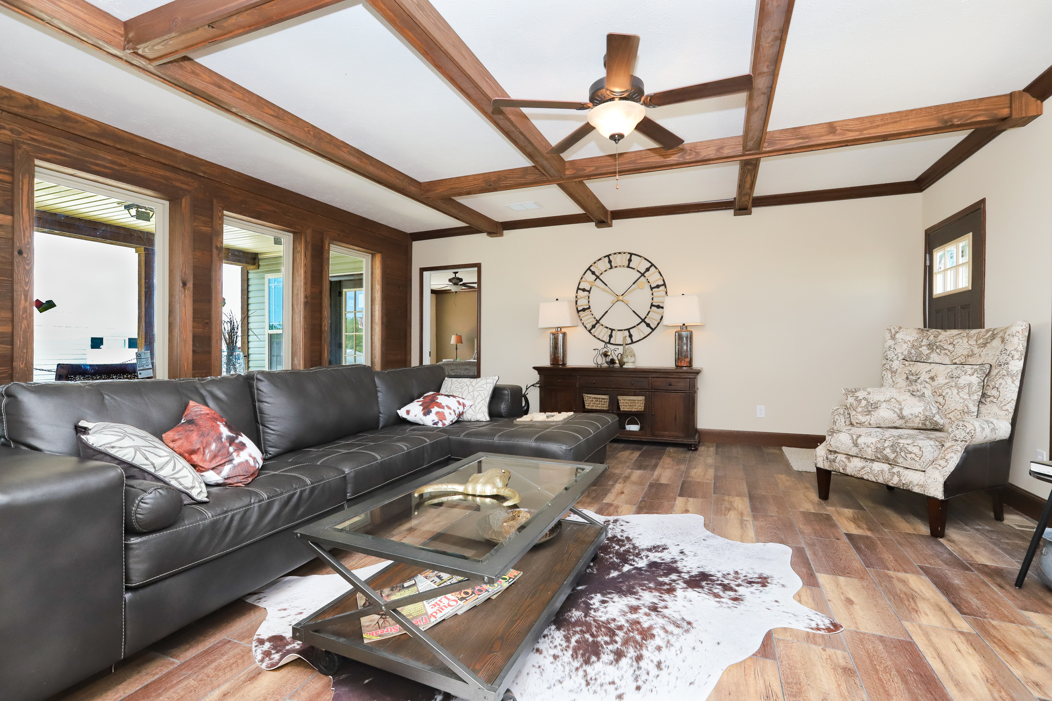 Spacious living room with a dark leather sectional sofa, glass coffee table, and patterned armchair. Wooden beams, large wall clock, cozy and inviting.