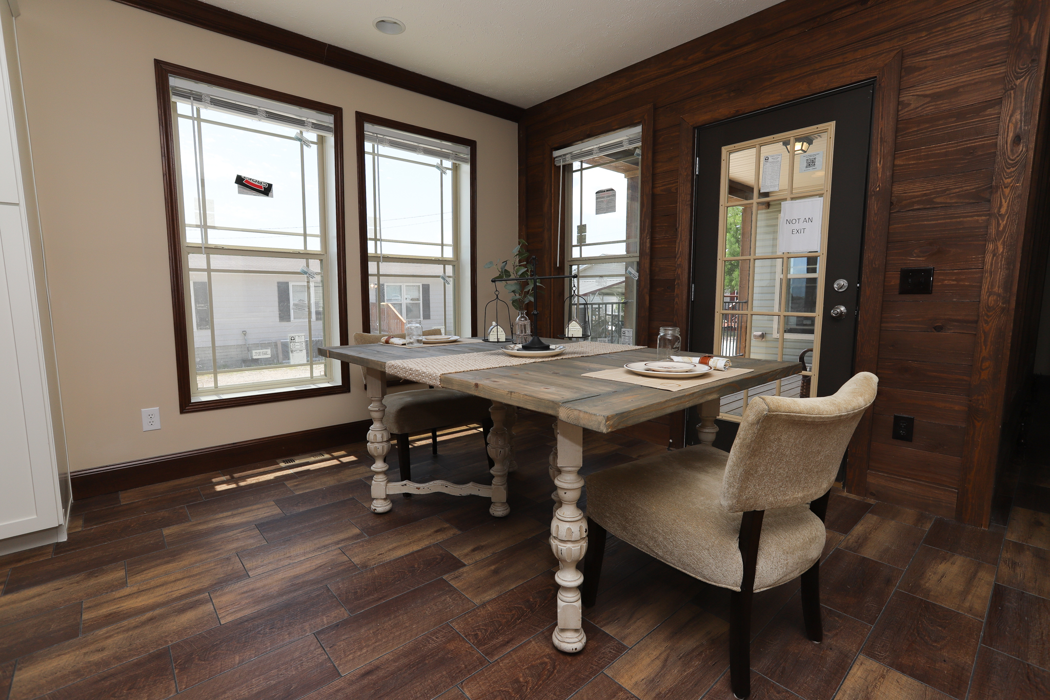 Dining area with a rustic wooden table set for two, beige upholstered chairs, and large windows with abundant natural light. Warm, inviting ambiance.