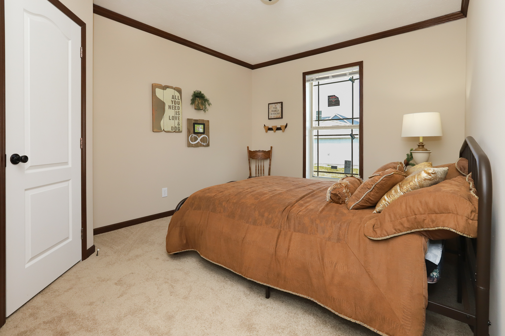 Warm-toned bedroom with brown bedding, a single window, and a lamp on one side. Wall decor includes a motivational sign and a small wreath. Cozy and inviting atmosphere.