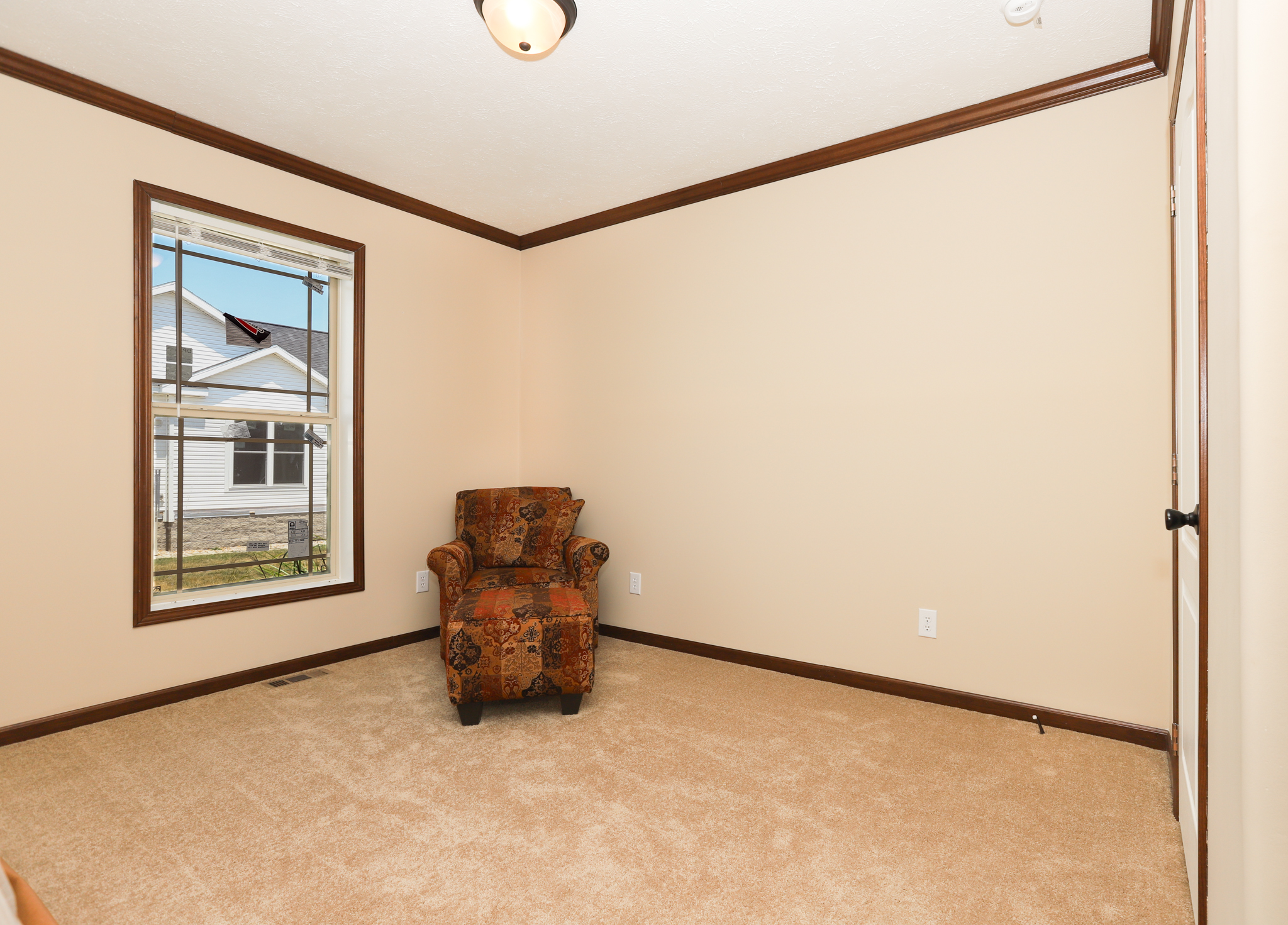A small, beige room with a single patterned armchair by a window. The window shows a view of a suburban house. The room is carpeted, conveying simplicity.