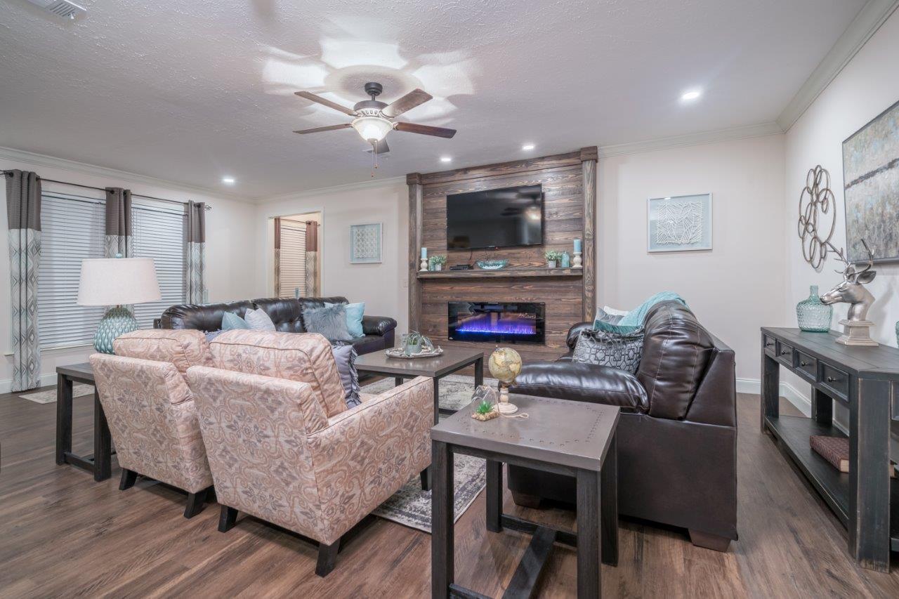 Stylish living room with a ceiling fan, brown leather sofas, patterned armchairs, and a wooden accent wall with a mounted TV and electric fireplace. Cozy ambiance.