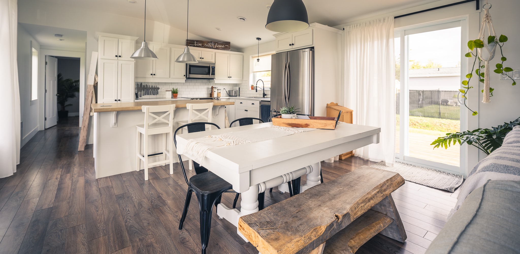 A modern kitchen and dining area with white cabinets, stainless steel appliances, and a large wooden table. Natural light streams through glass doors.
