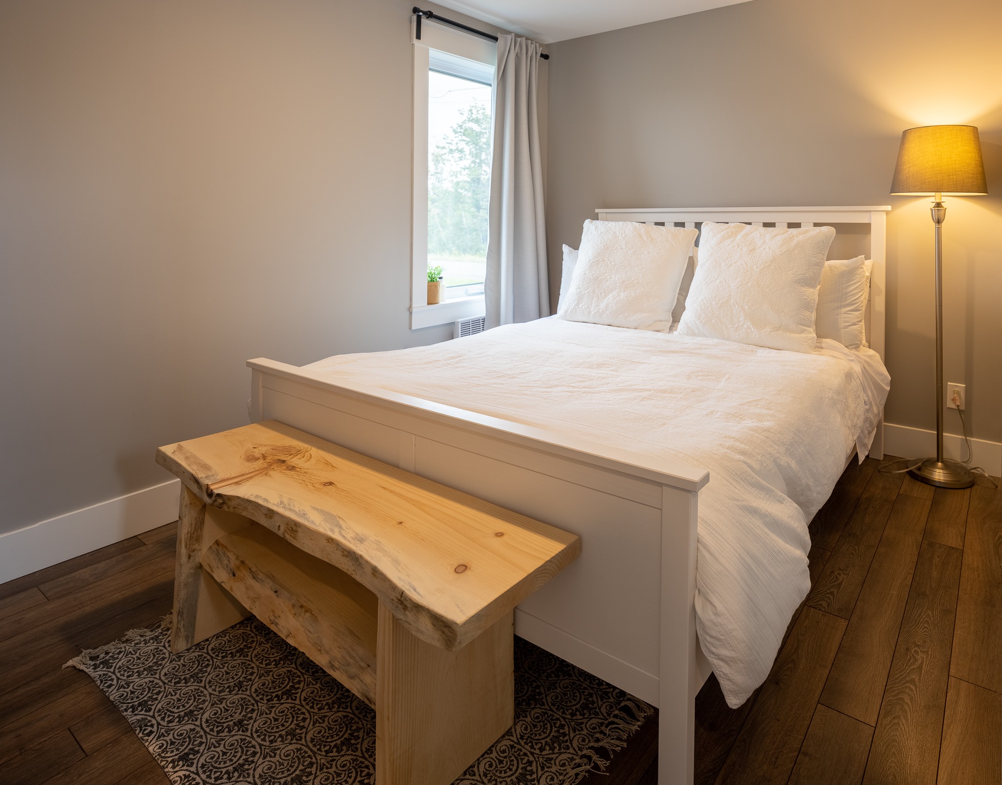 Cozy bedroom with a white bed, fluffy pillows, wooden bench, soft lighting from a lamp, gray walls, and a window with curtains. Calm and inviting.