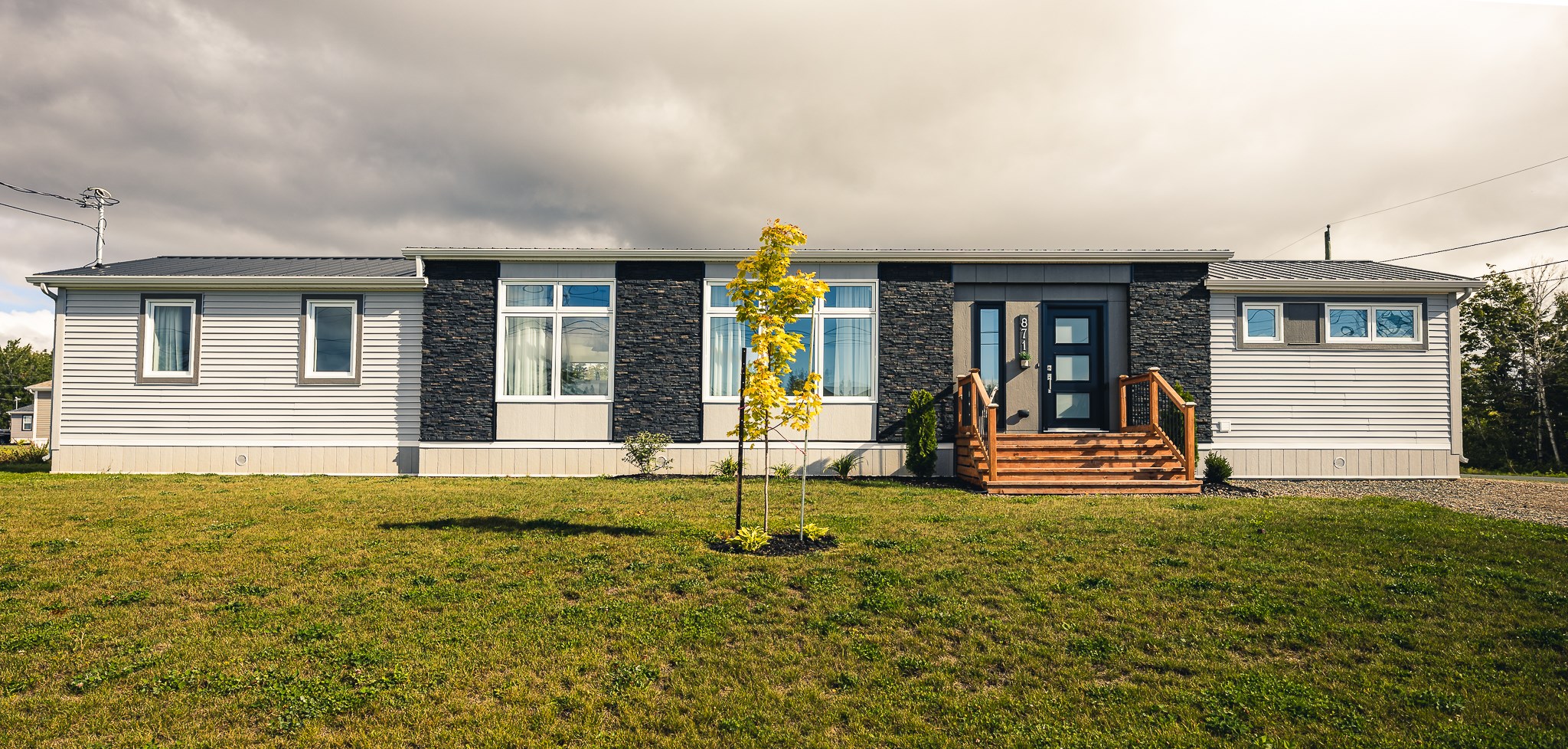 A modern, single-story house with light siding and dark stone accents. Wooden steps lead to the front door. A small tree stands on the green lawn.