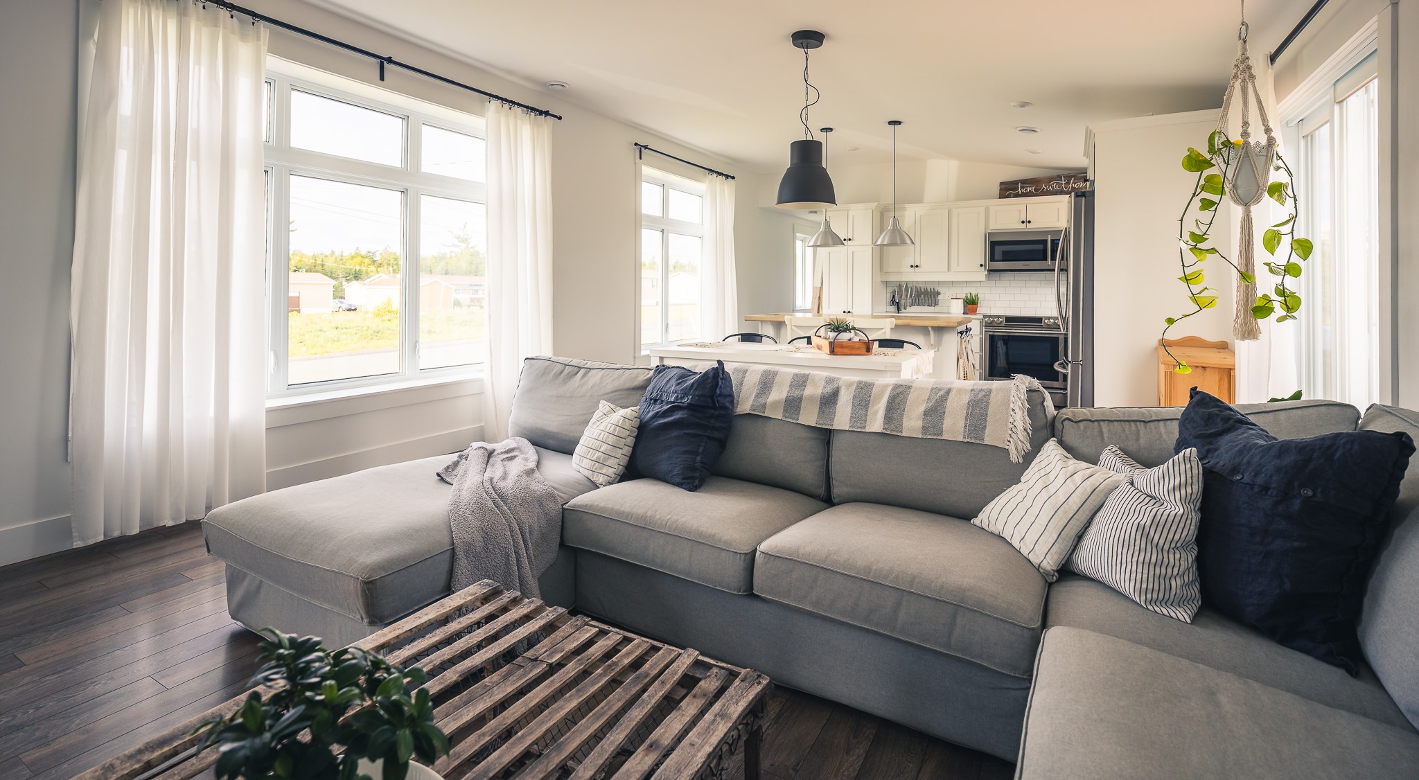A cozy living room with a plush gray sectional sofa adorned with navy and striped pillows. Sunlight pours through large windows, illuminating the warm wooden floor and a rustic wooden coffee table. A hanging plant adds a touch of greenery, and a modern kitchen is visible in the background, conveying a welcoming and serene atmosphere.