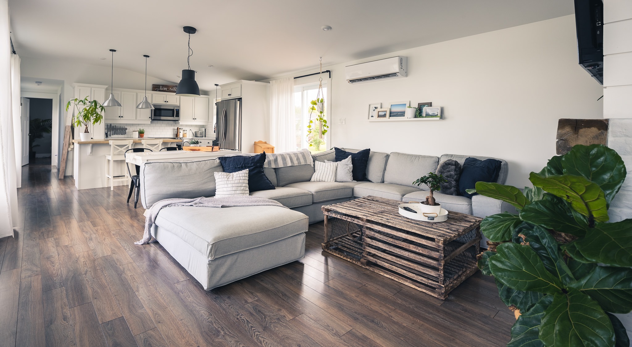 Modern living room with a spacious gray sectional sofa, wooden coffee table, and lush green plants. Open kitchen with pendant lights in the background. Cozy and inviting atmosphere.