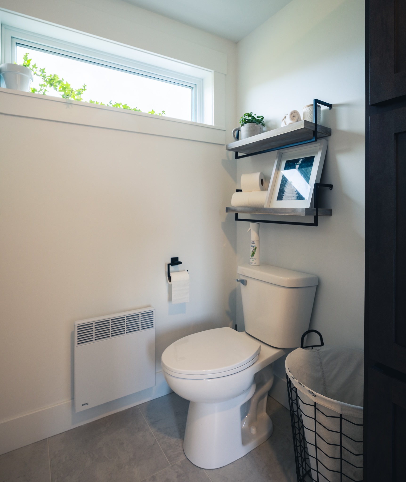 Compact bathroom with white walls, featuring a toilet, metal shelf with plants, and rolled towels. Soft natural light from a small window adds warmth.