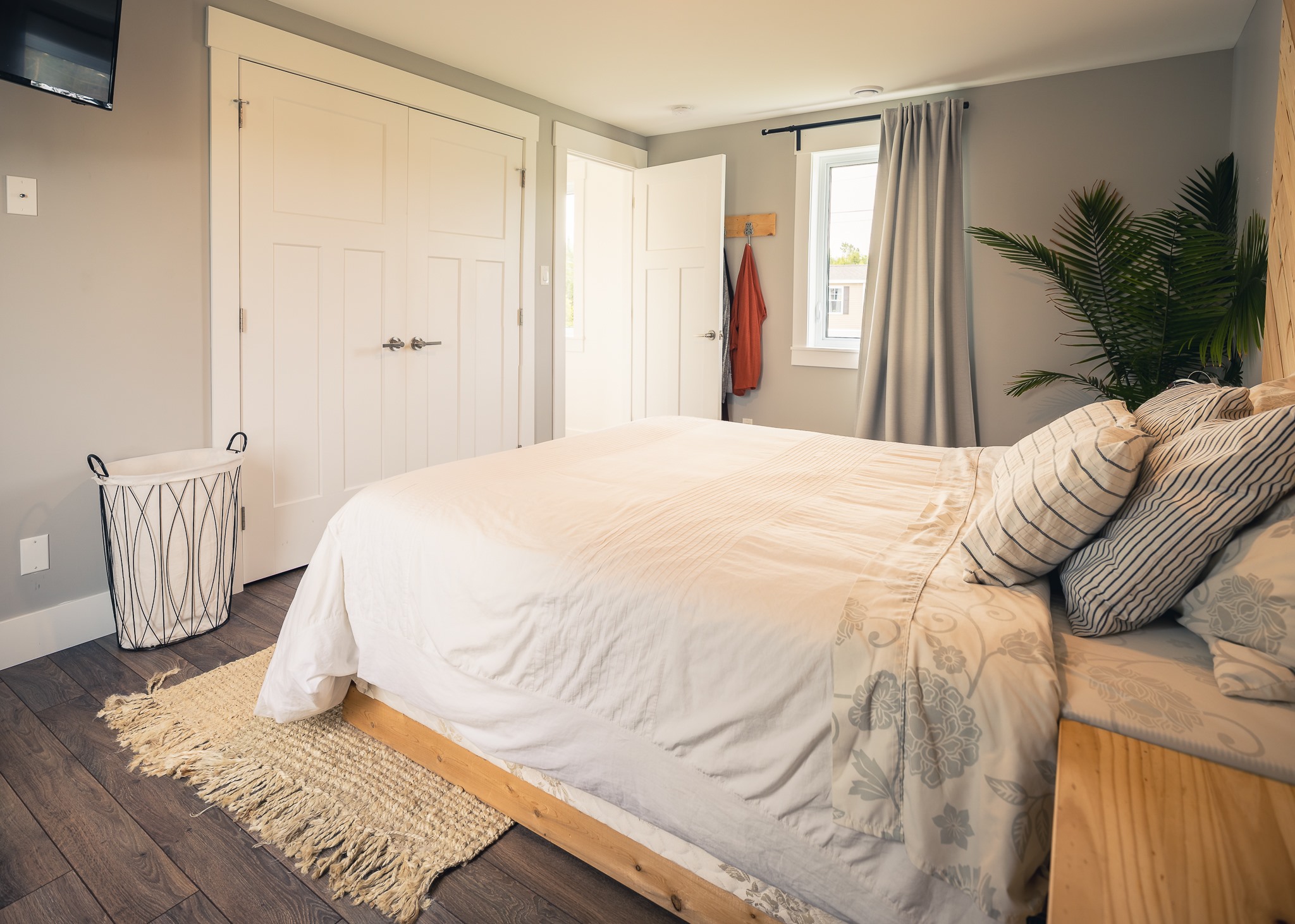 Cozy bedroom with neutral tones, featuring a neatly made bed, decorative pillows, and potted plant. Sunlight filters through a window with gray curtains.