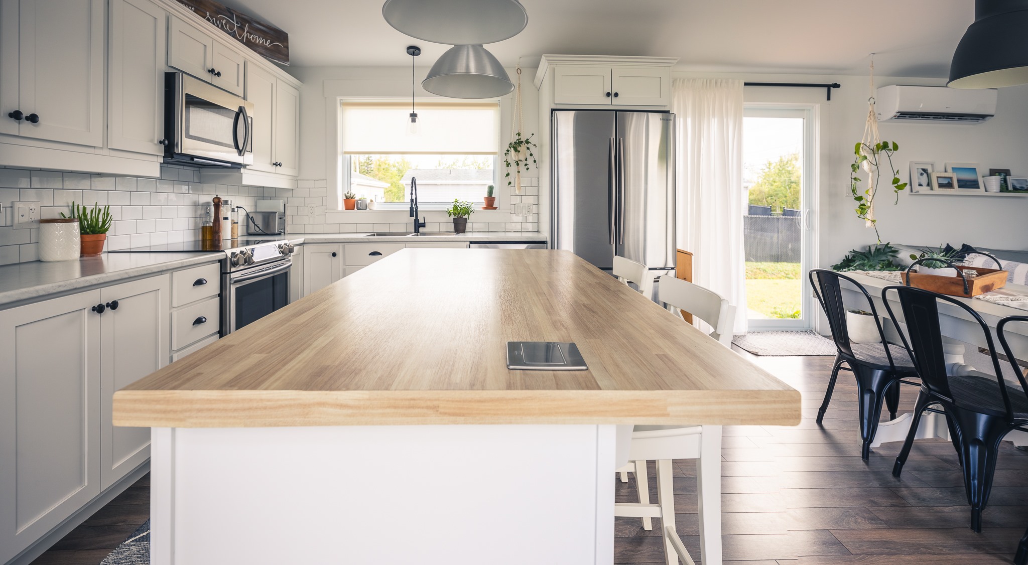 Bright, modern kitchen with a wooden island, white cabinets, and stainless steel appliances. Sunlight streams through a large window and glass door.