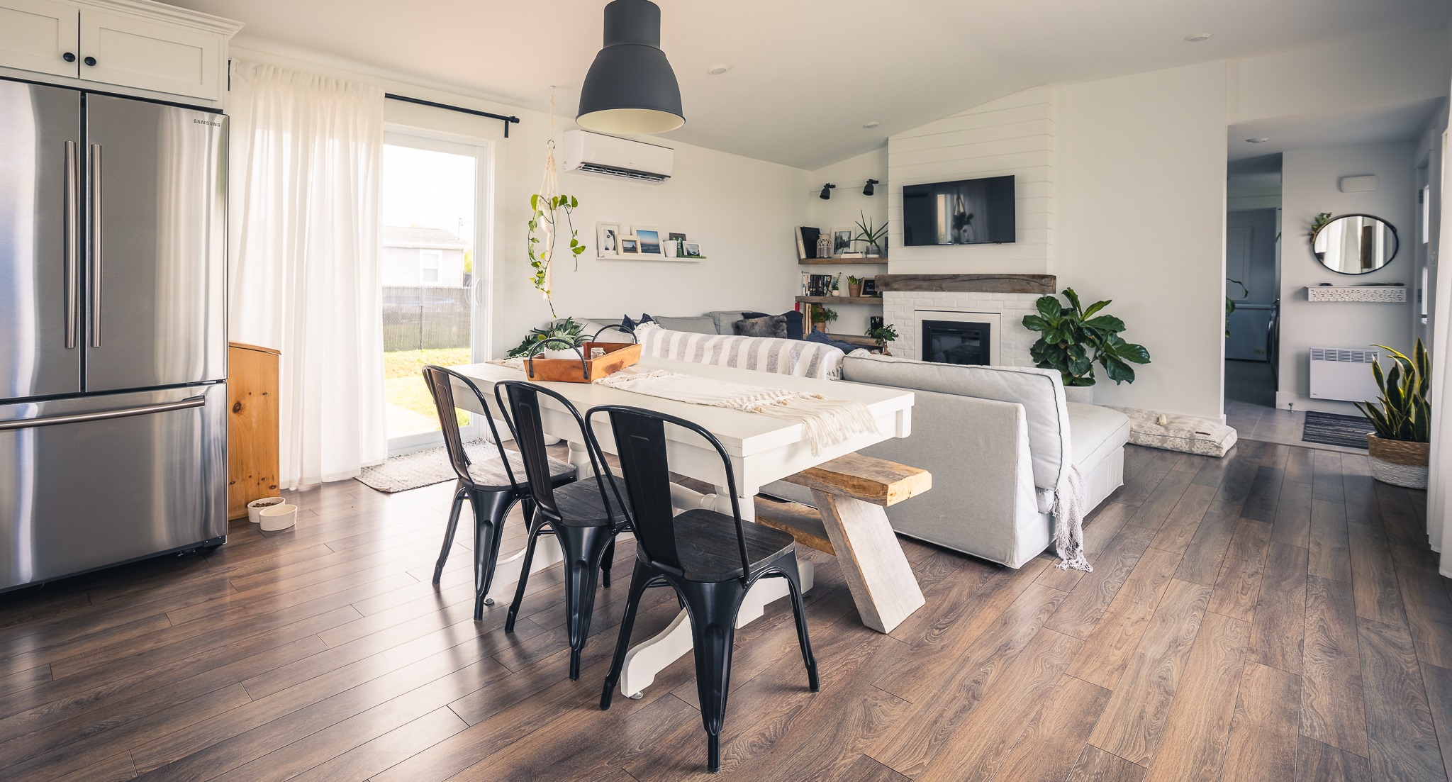 A modern living-dining area with wooden floors, a gray sofa, white table with black chairs, stainless steel fridge, plants, and a cozy ambiance.