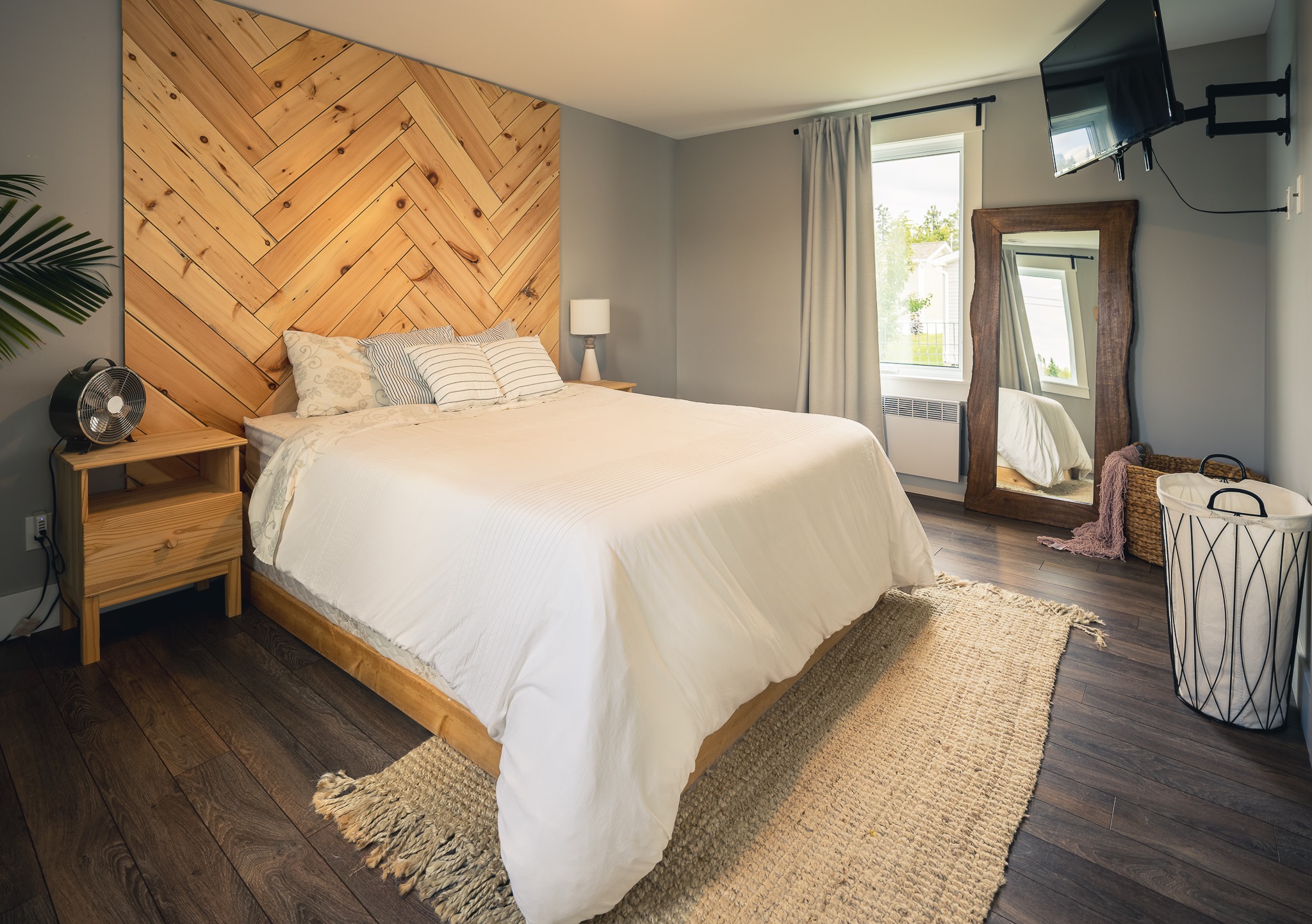 Cozy bedroom with a chevron wood accent wall, a neatly made bed with white linens, wooden nightstand, wall-mounted TV, large mirror, and basket. Warm ambiance.