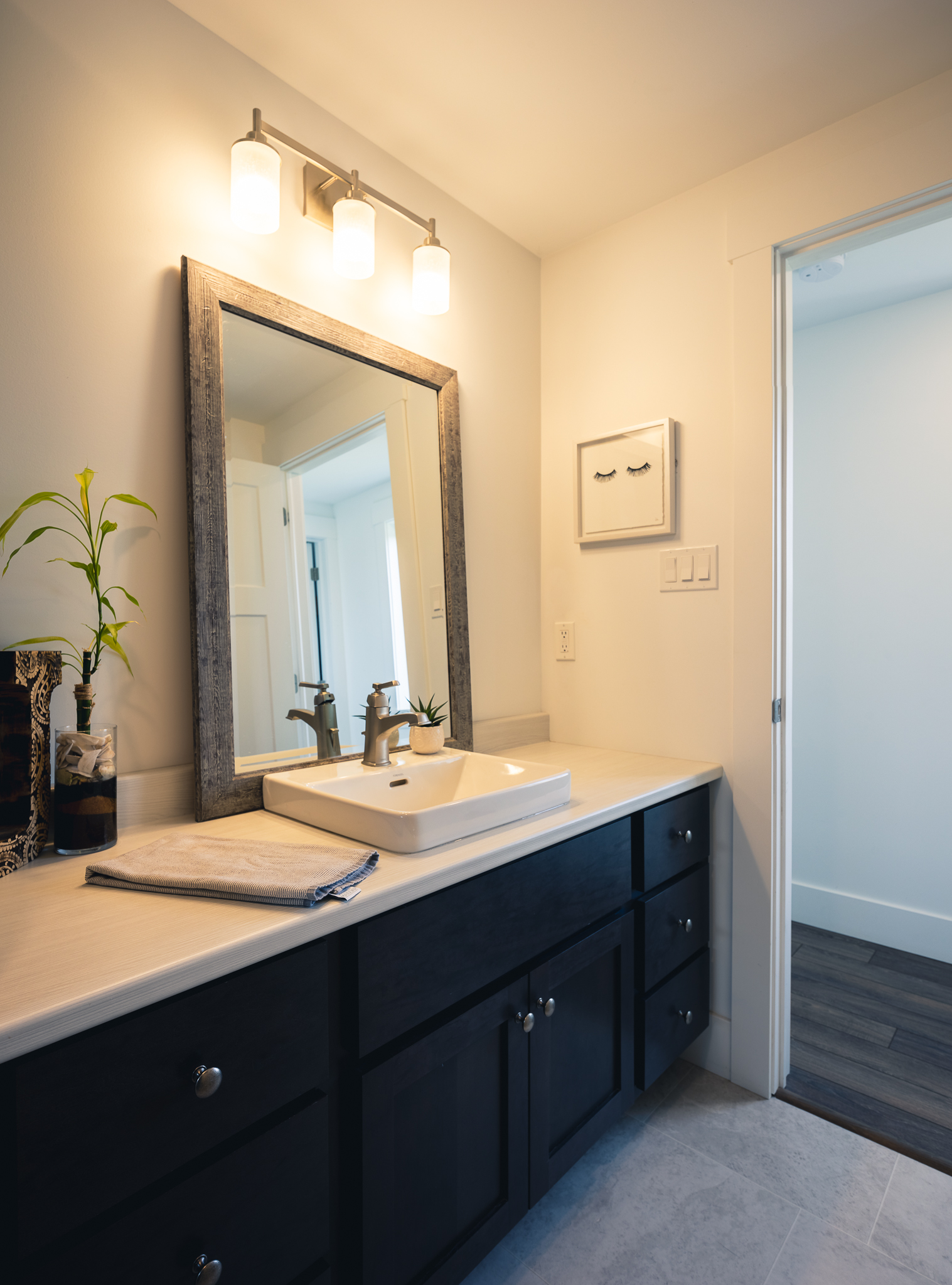 Modern bathroom with a sleek countertop and square sink, framed mirror above, bright vanity lights, and a small plant for decor. Calm and tidy ambiance.