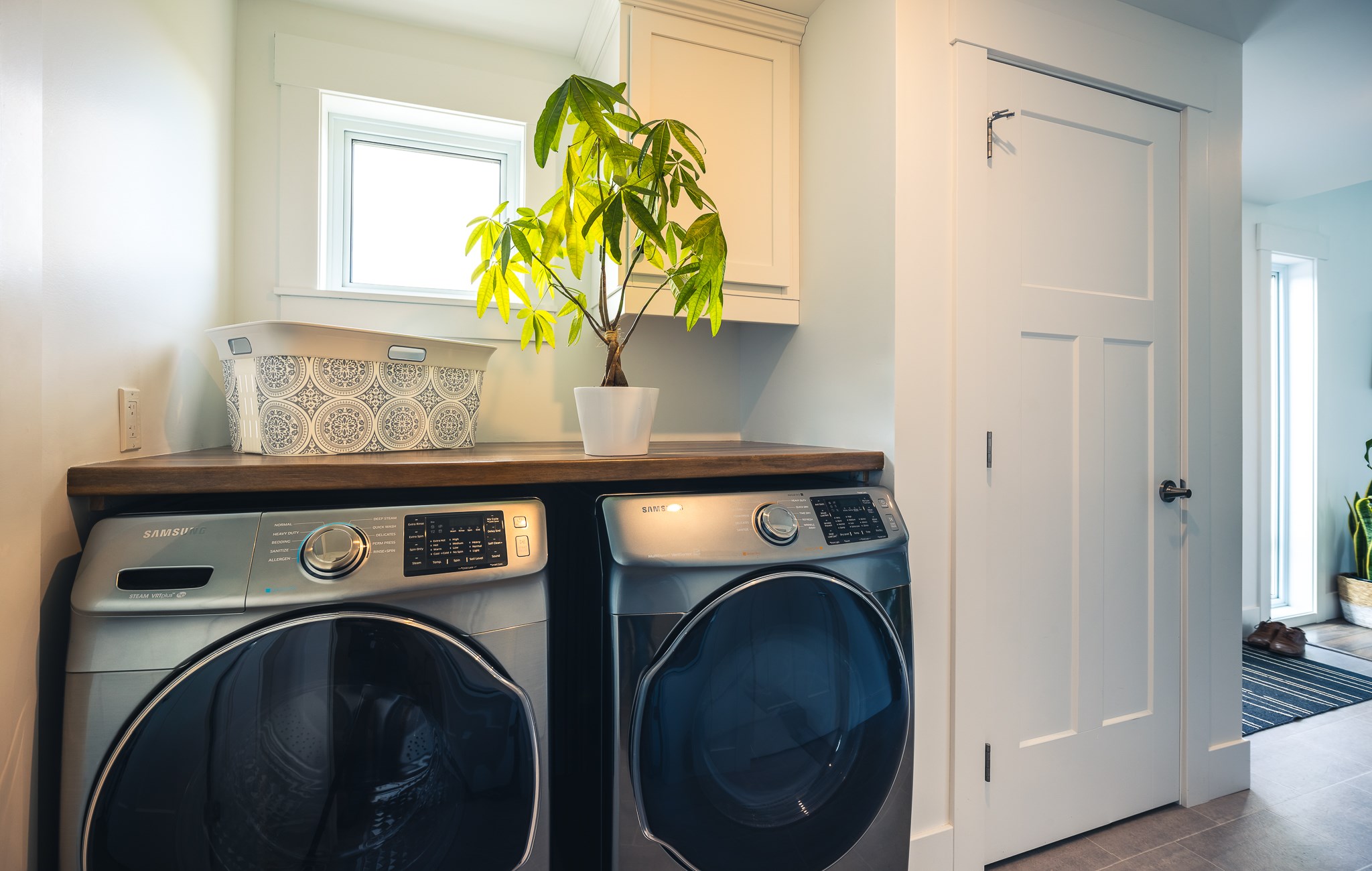 A modern laundry room with a silver washer and dryer set topped with a wooden counter. A potted plant and a patterned basket add a fresh touch.