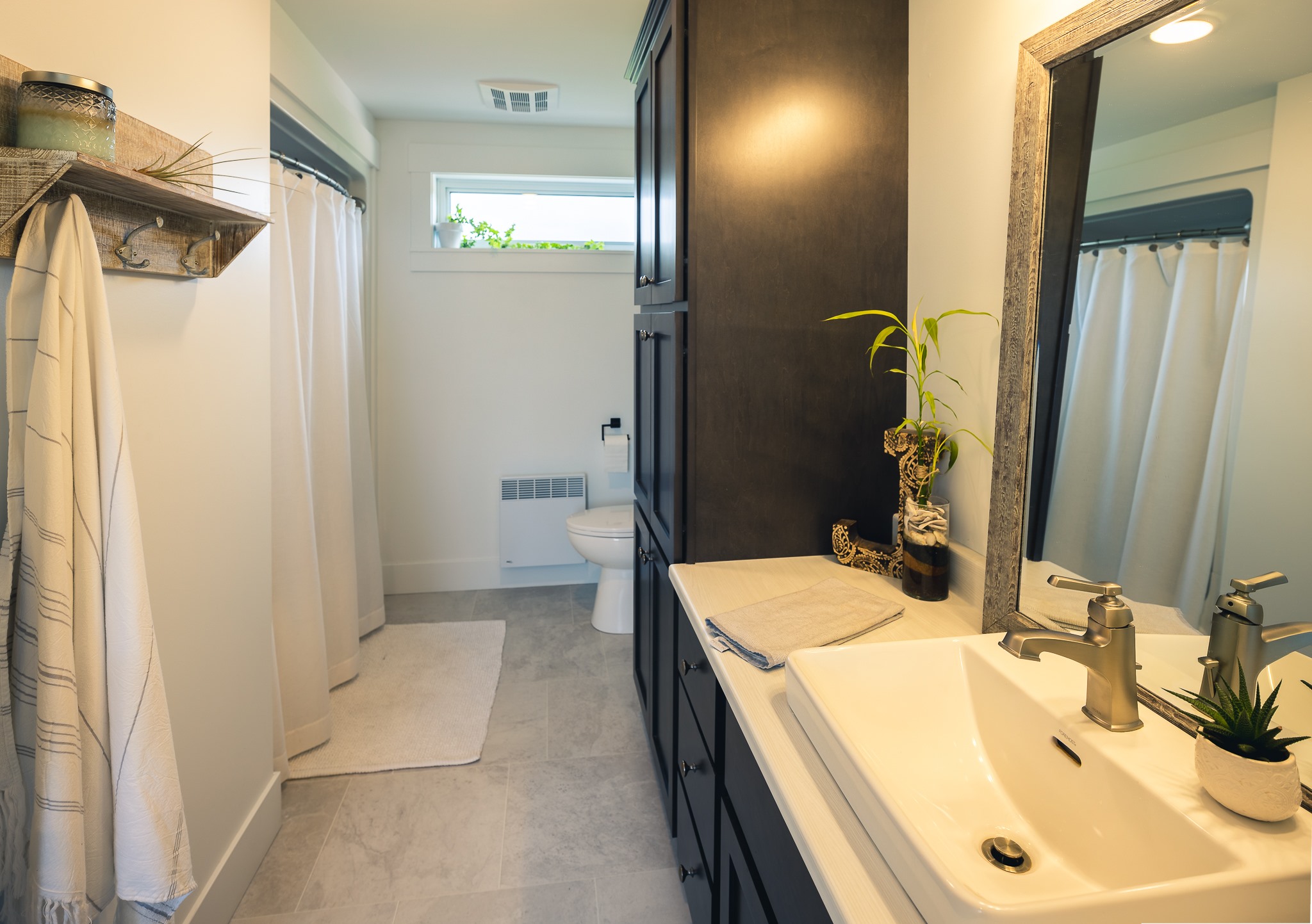Modern bathroom with a white sink, dark wood cabinetry, and a large mirror. Curtains, potted plants, and soft lighting create a calm atmosphere.
