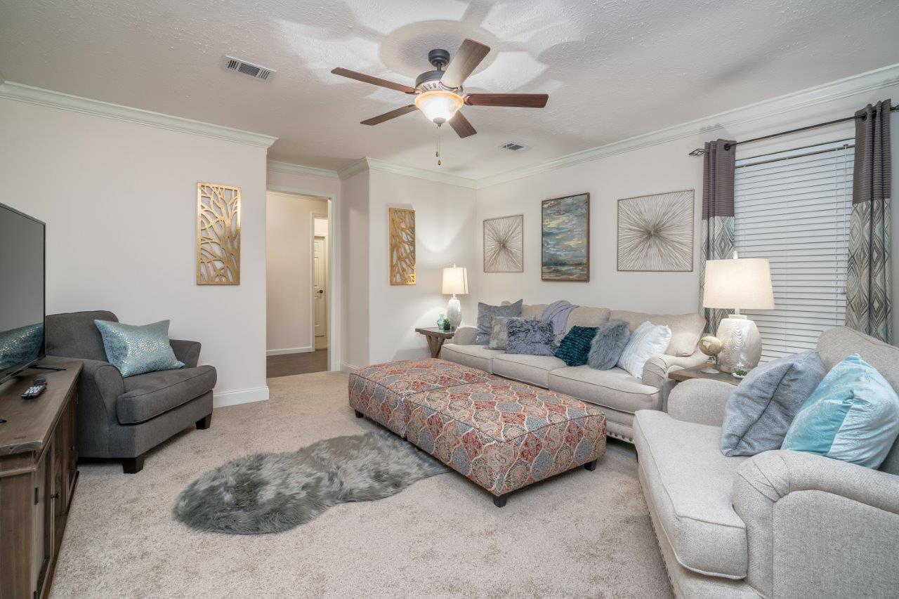 Cozy living room with a cream sectional sofa adorned with blue and gray cushions, a patterned ottoman, and artistic wall decor. Warm lighting and beige carpet create a relaxed atmosphere.