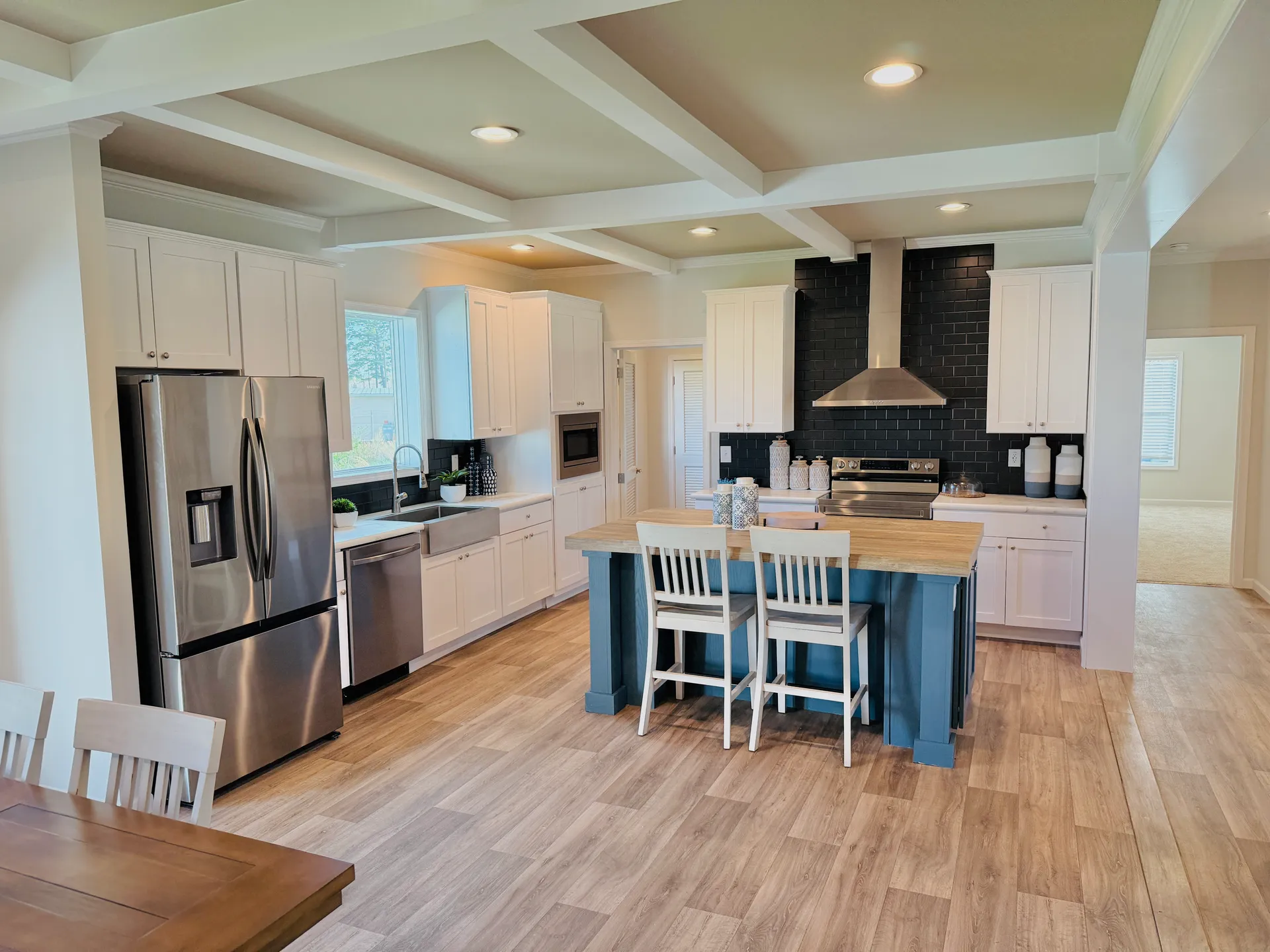 Modern kitchen with white cabinets, a stainless steel fridge, and a central island with blue base. Wooden floor, black tile backsplash, and coffered ceiling. Bright and inviting.
