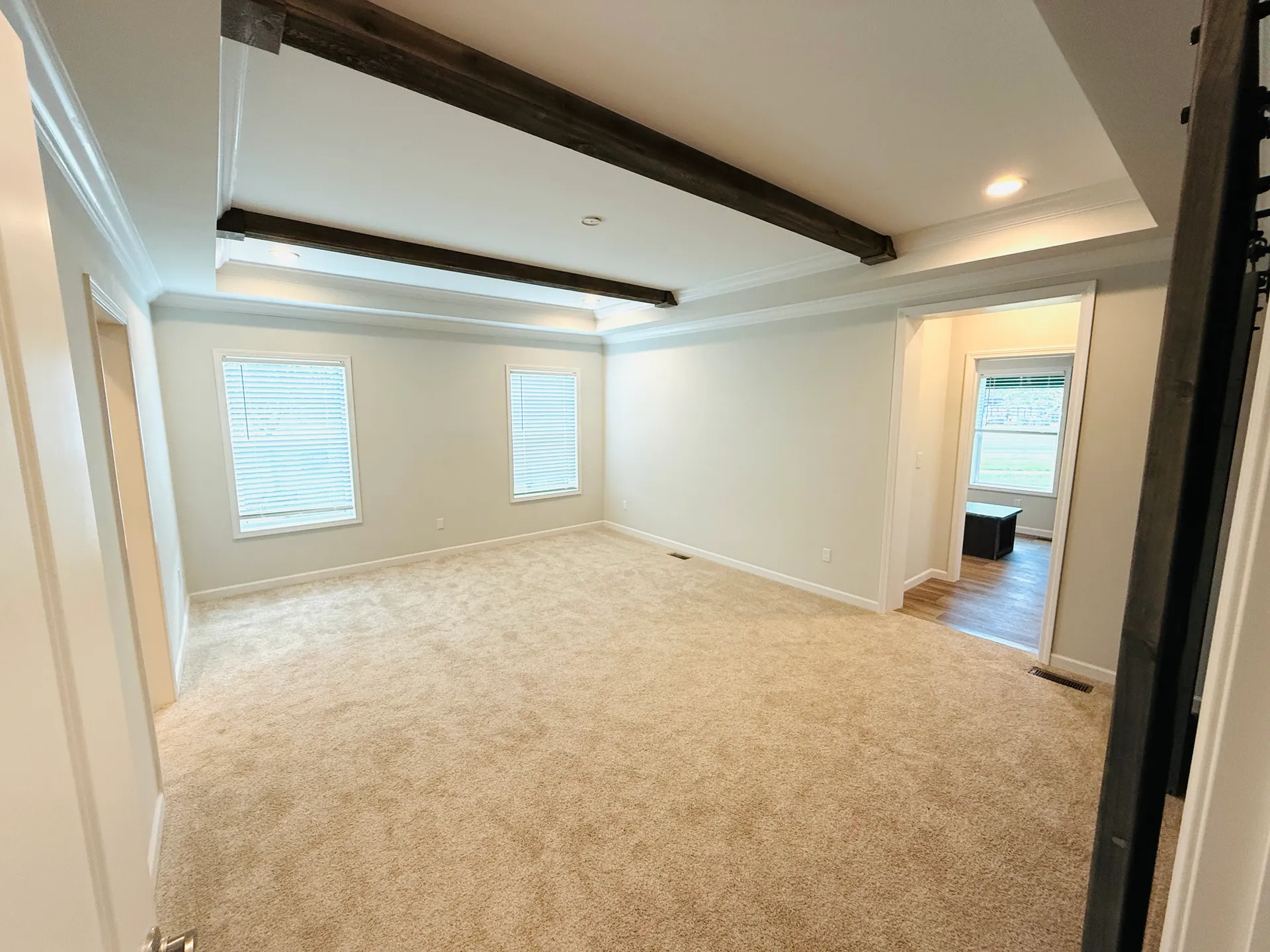 Spacious room with beige carpet, bright walls, and dark wood ceiling beams. Two windows with blinds allow light in, creating a calm, inviting space.