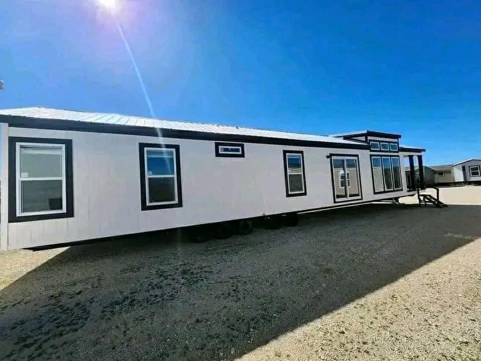 A modern white mobile home with dark trim sits on gravel under a bright blue sky. Sunlight creates a soft glow, conveying a sense of openness and tranquility.