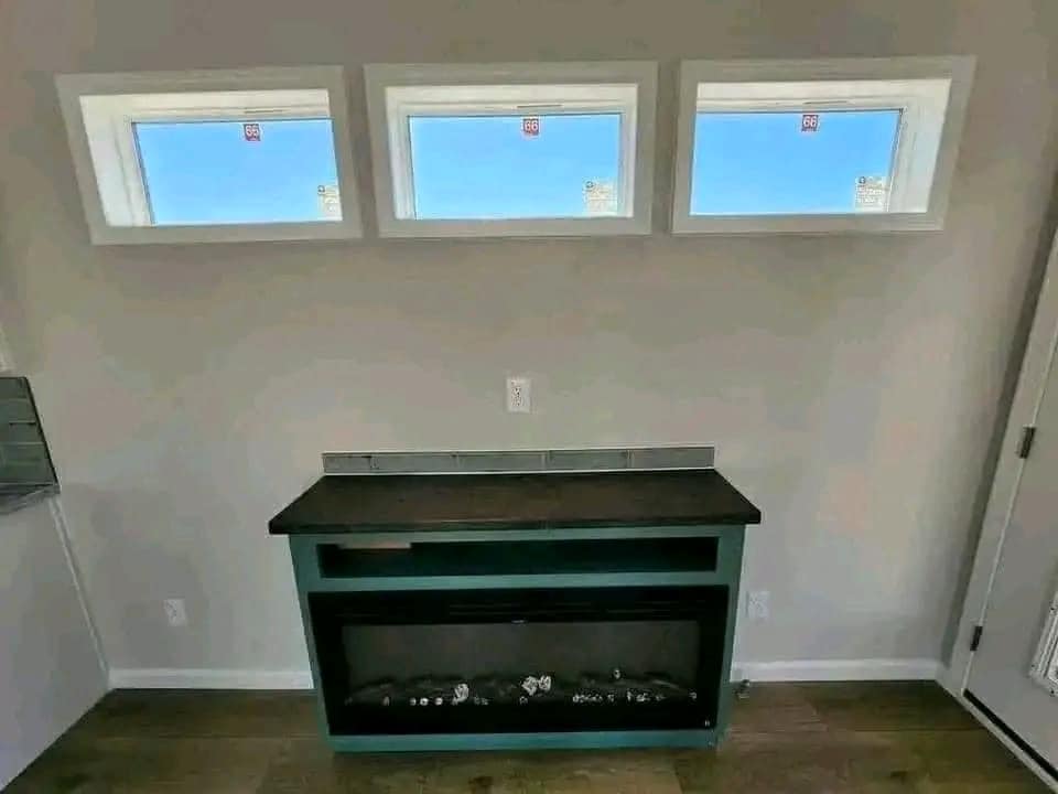 A modern room features three small rectangular windows with clear blue skies. Below is an electric fireplace with a black countertop, creating a sleek ambiance.