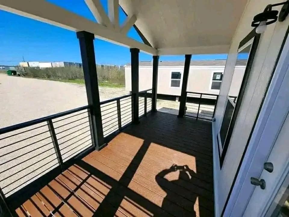 Covered porch with wooden flooring and black railings, casting shadows under bright sunlight. A person’s shadow appears, capturing the view. Background shows a clear blue sky and neighboring buildings.