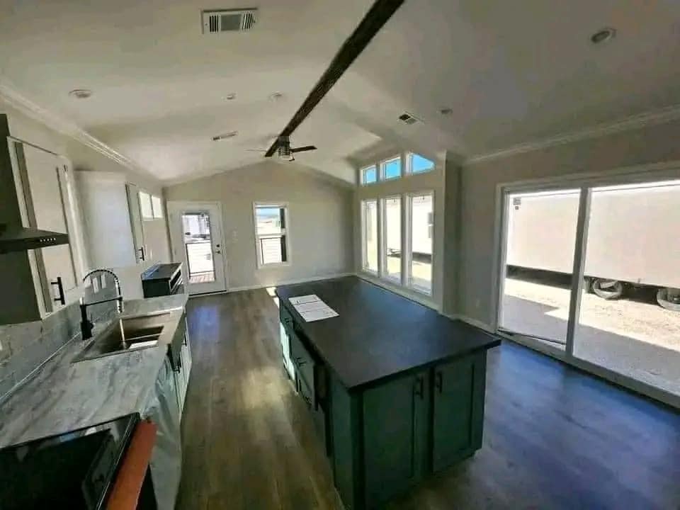 Modern kitchen interior with high ceilings, wood flooring, a central island with black countertop, white cabinets, and large windows providing natural light.