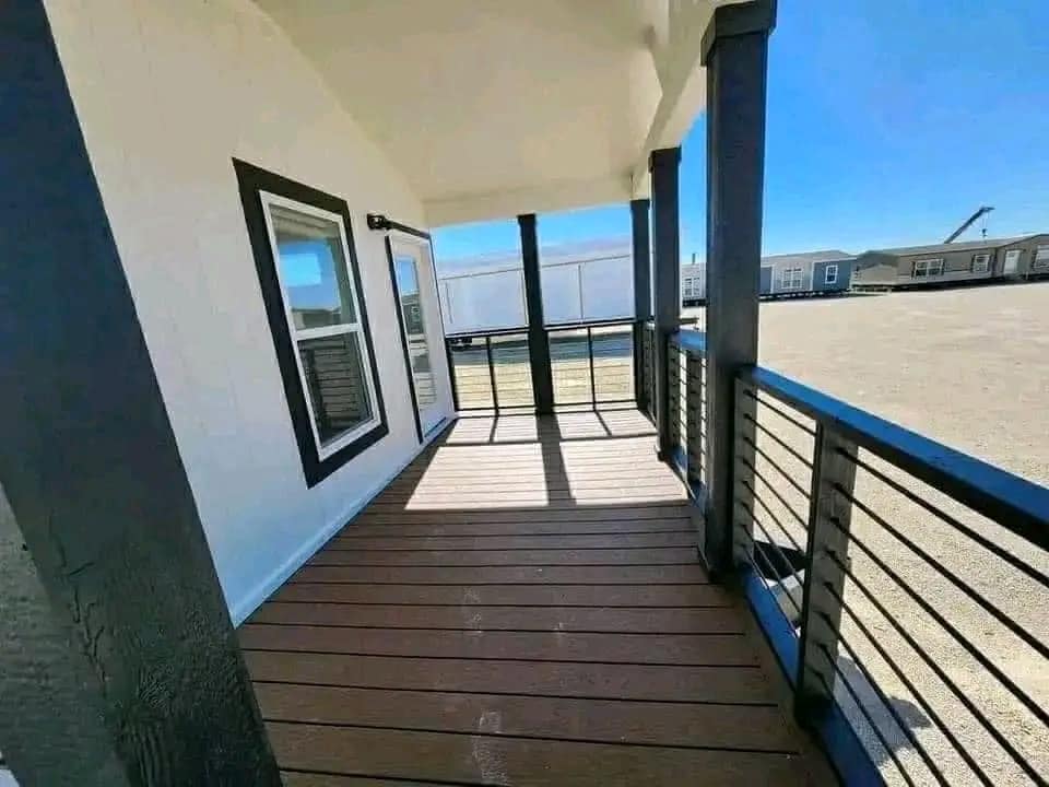 Covered porch with wooden flooring and black railing, attached to a white house with black-trimmed windows. Sunlight casts strong shadows, creating a serene and inviting atmosphere.