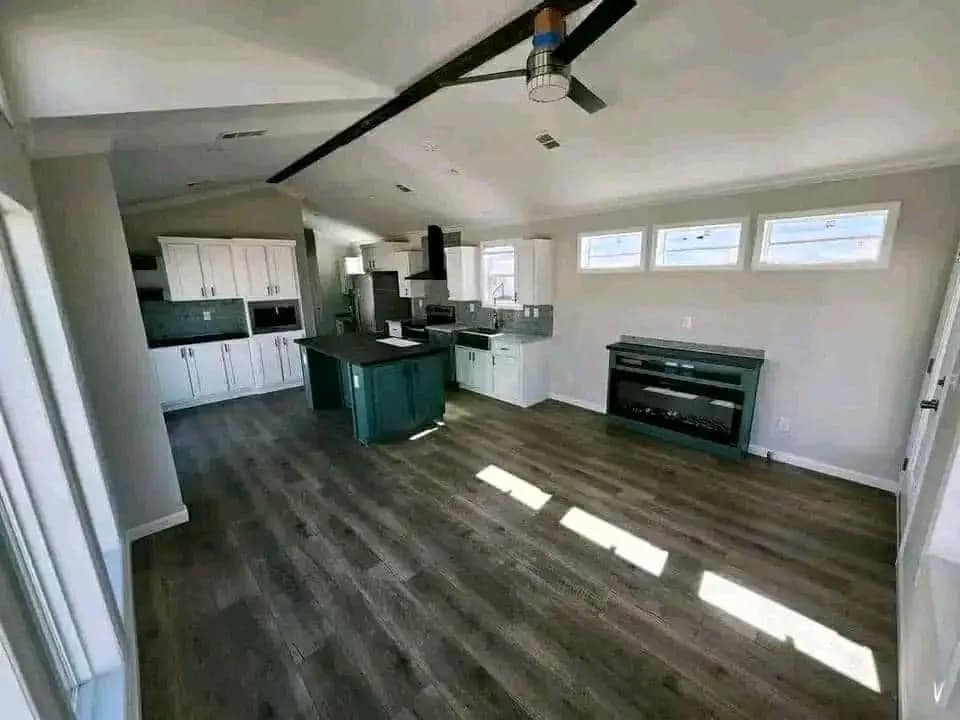 Spacious modern kitchen with white cabinets, dark wood floor, teal island, ceiling fan, and large windows casting sunlight, creating an airy feel.