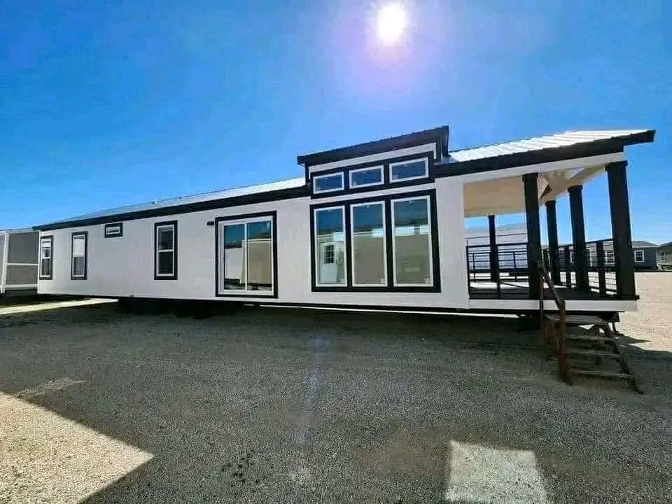 Modern tiny house with large windows and a covered porch on a sunny day. The blue sky and shadows enhance the home's sleek and inviting look.
