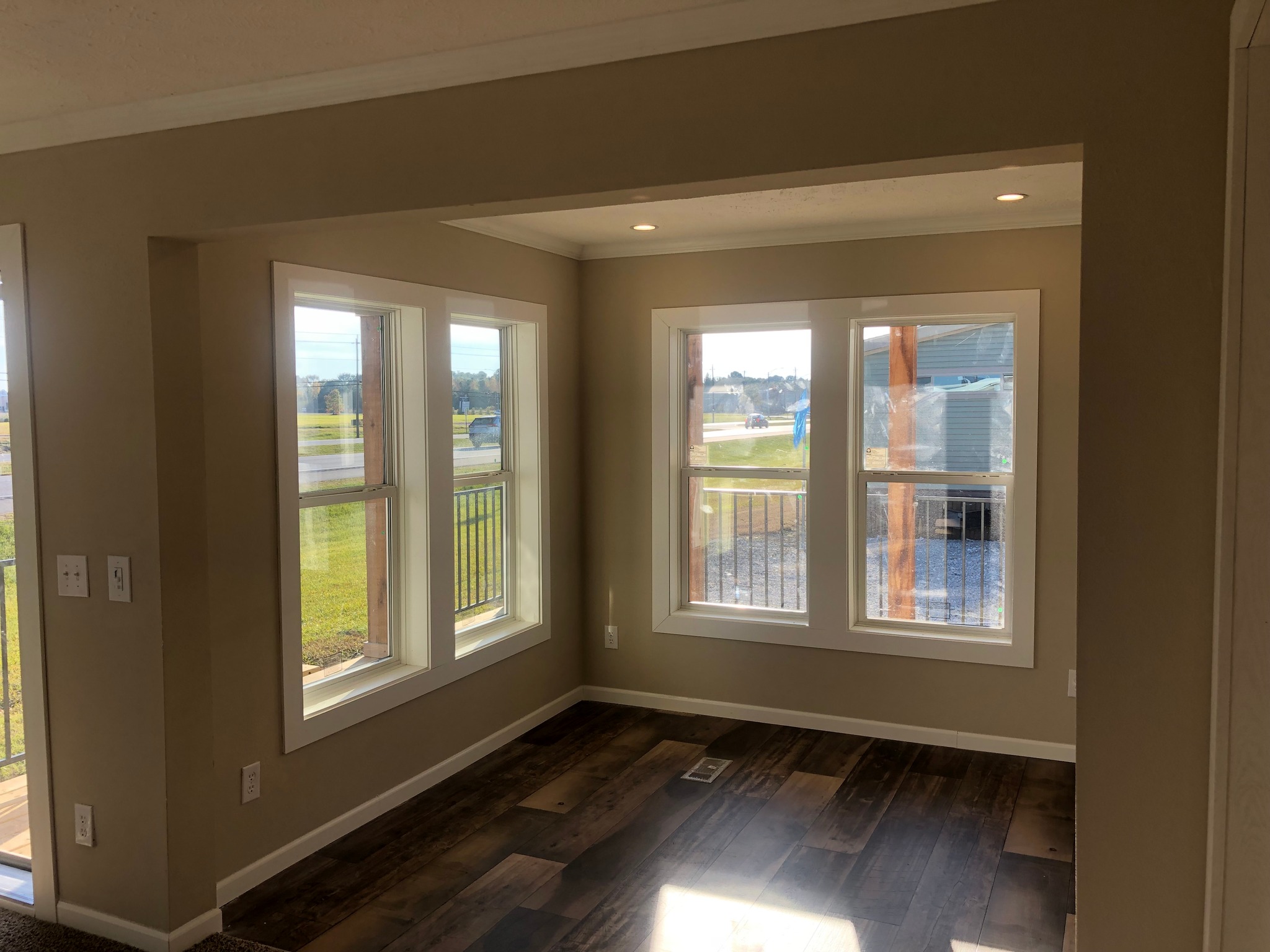 Bright room with brown wooden floor and large corner windows showcasing a scenic view. Sunlight streams through, creating a warm, inviting ambiance.