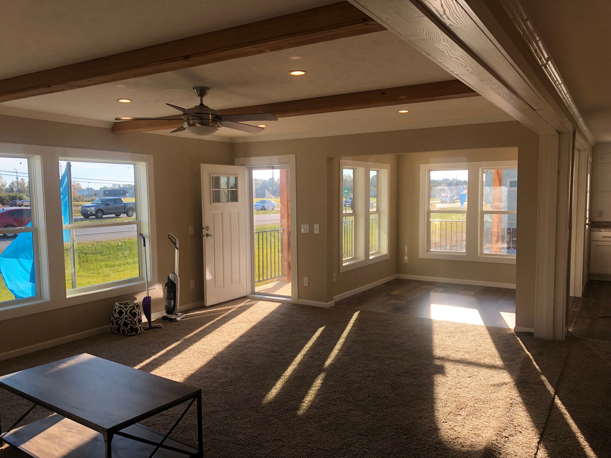 Bright living room with large windows, open door, ceiling fan, and sunlight streaming in. The space is airy with beige walls, carpet, and a small table.