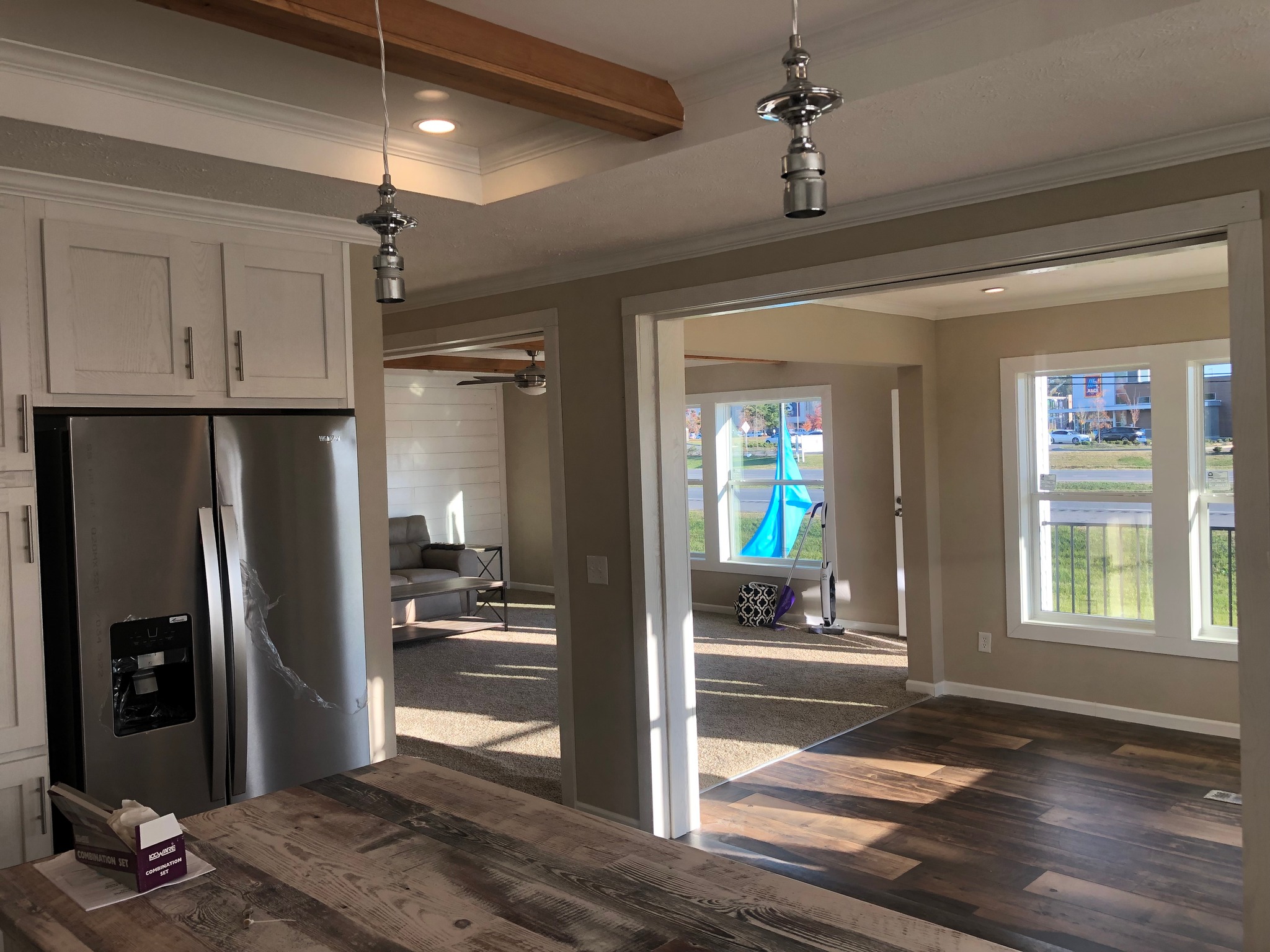 Open modern kitchen and living area with wood and white decor. Sunlight streams through large windows, creating a cozy, inviting atmosphere. A stainless steel fridge stands near a rustic table.