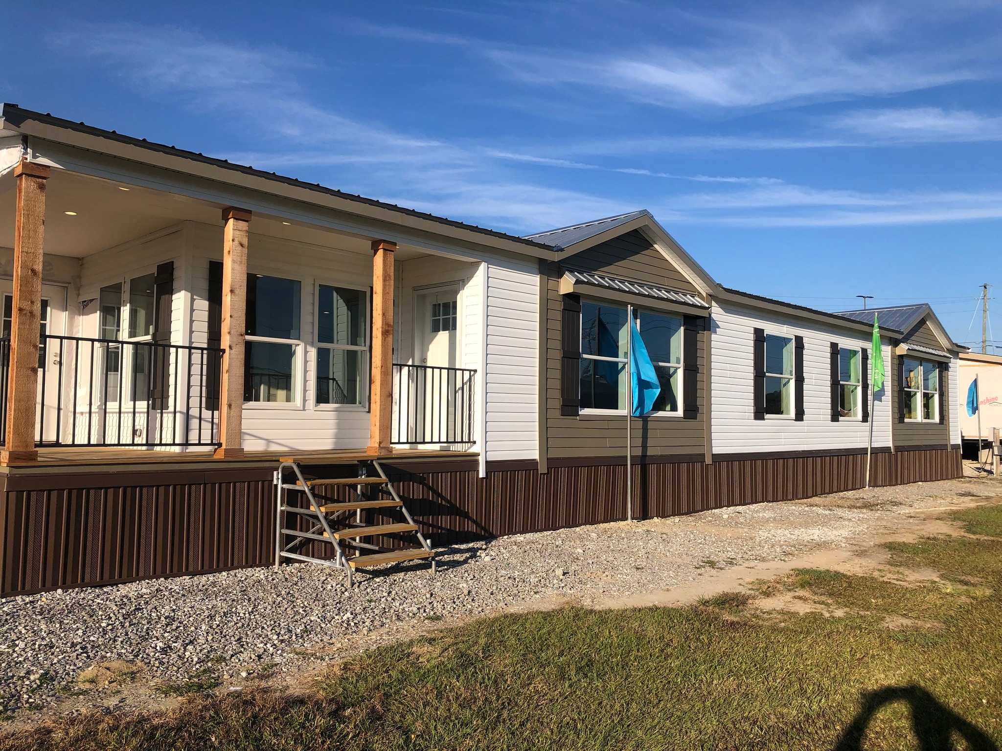 A modern prefabricated home with a porch, metal railing, and wooden pillars is set on a gravel plot. Blue sky and grassy foreground convey a serene setting.