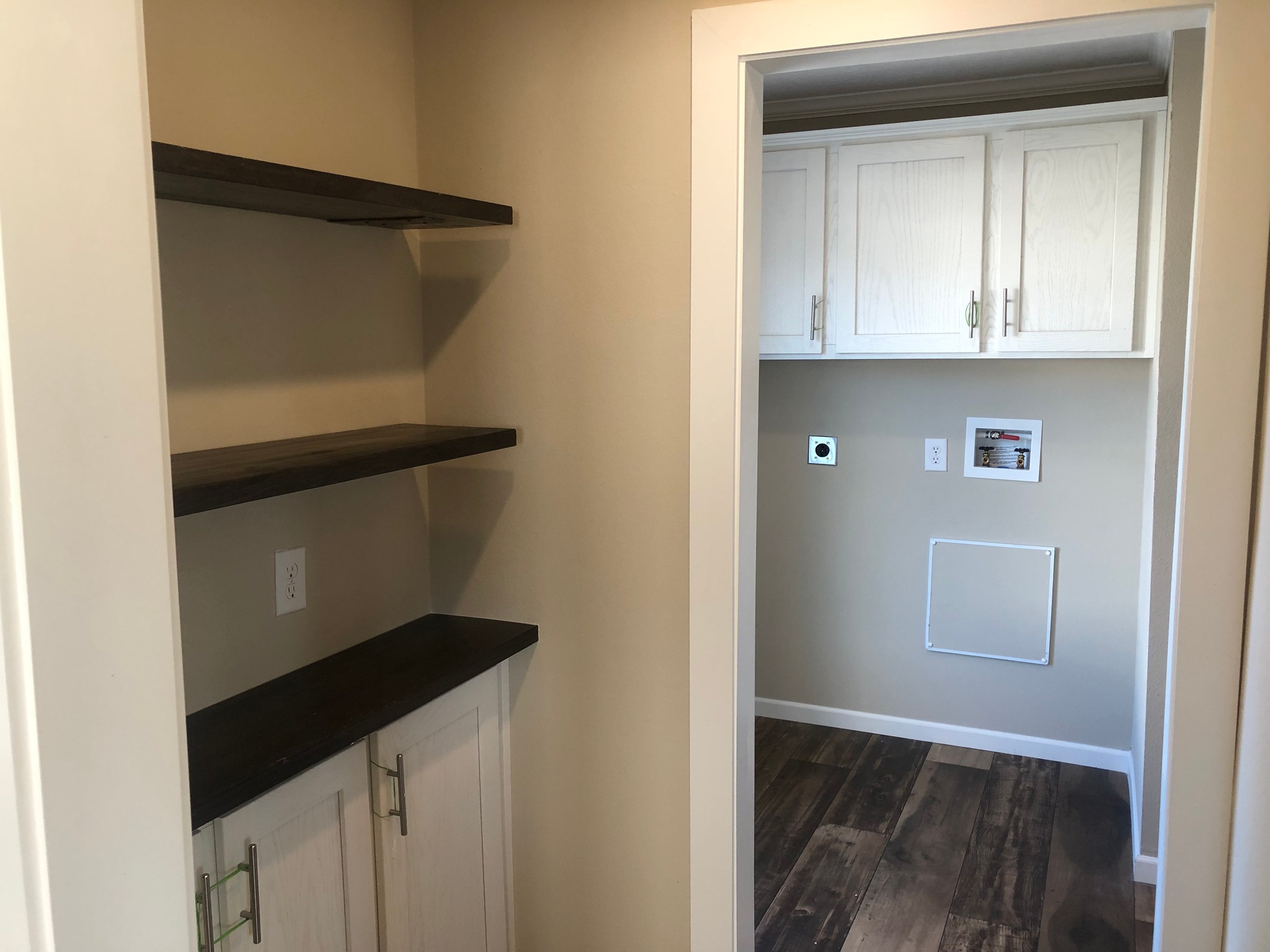 A small, beige mudroom with dark wooden shelves on the left and white cabinets opposite. The floor features dark wood planks, creating a cozy, organized space.