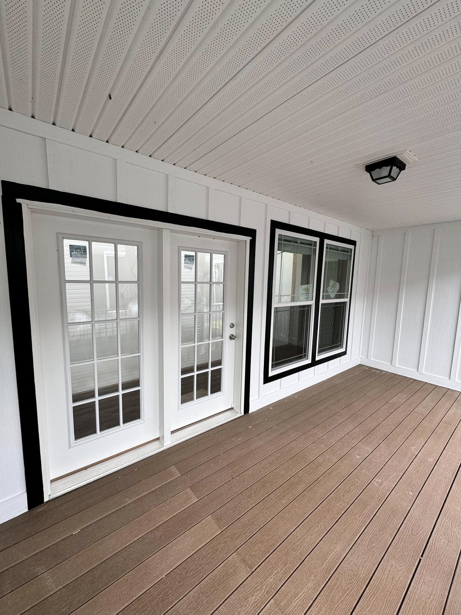 Enclosed porch with white walls and a brown floor features double glass doors and large windows framed in black. A ceiling light adds a modern touch.